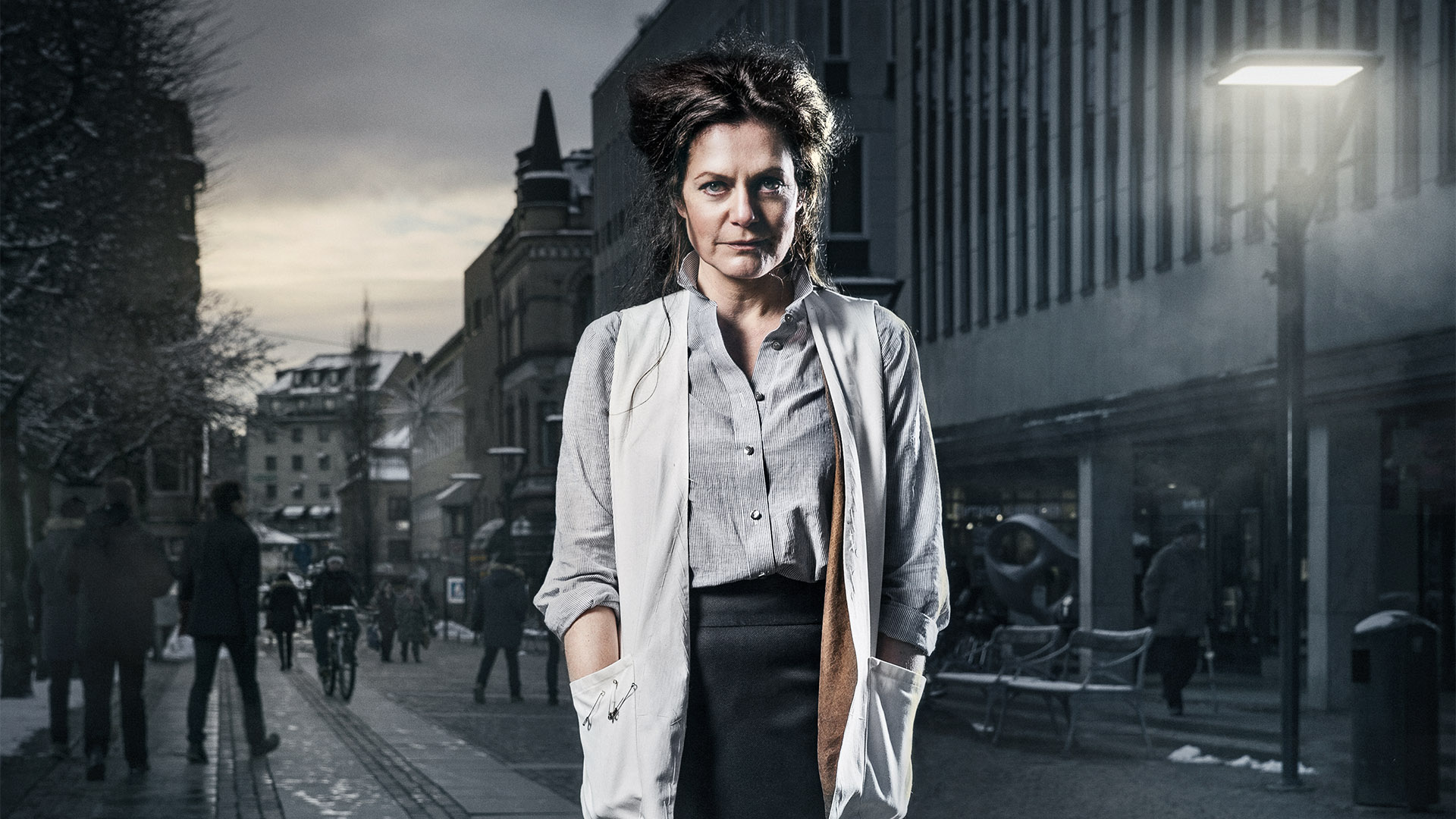 Klickbar bild Skandal. En kvinna vänd rakt mot kameran framför en grå vy över stora torget i Borås. Kvinnan har lockigt uppsatt hår, blårandig skjorta och vit väst med säkerhetsnålar fästa på ena fickan. 
