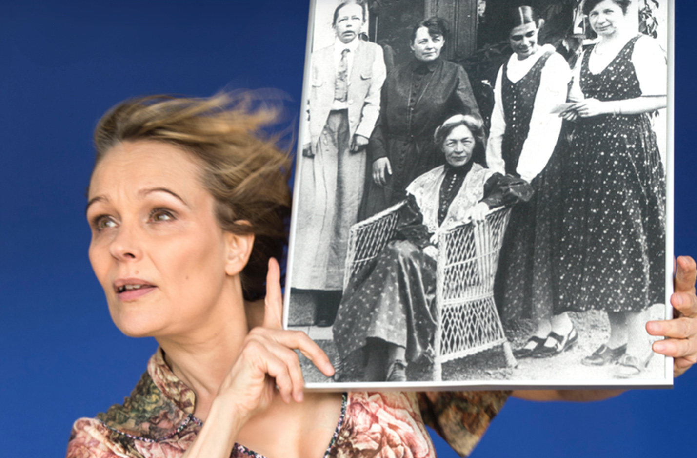 Klickbar bild Fogelstad. En skådespelare med långt blont hår som fladdrar i vinden håller upp ett svartvitt foto som föreställer kvinnorna som drev Fogelstad medborgarskola. 