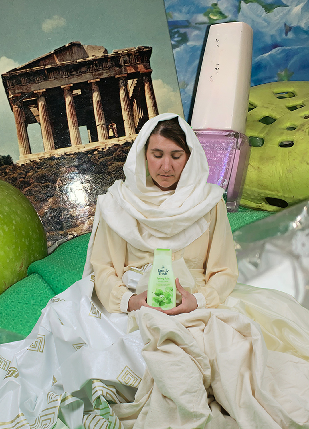 Fotokollage över olika föremål i ovanliga proportioner till varandra. En kvinna i ljus toga och sjal runt huvudet i förgrunden håller i en grön flaska med duschtvål. I barkunden ett enormt grönt äpple, ett hopskrynklat mintgrönt papper, ett vykort föreställande Akropolis, en gigantisk flaska med rosa nagellack och en starkt grön foppatoffla.  