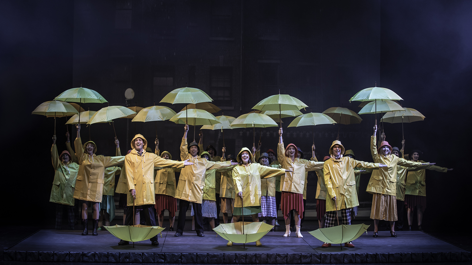 Stor ensemble i gula regnrockar med gula paraplyer på stora scenen.