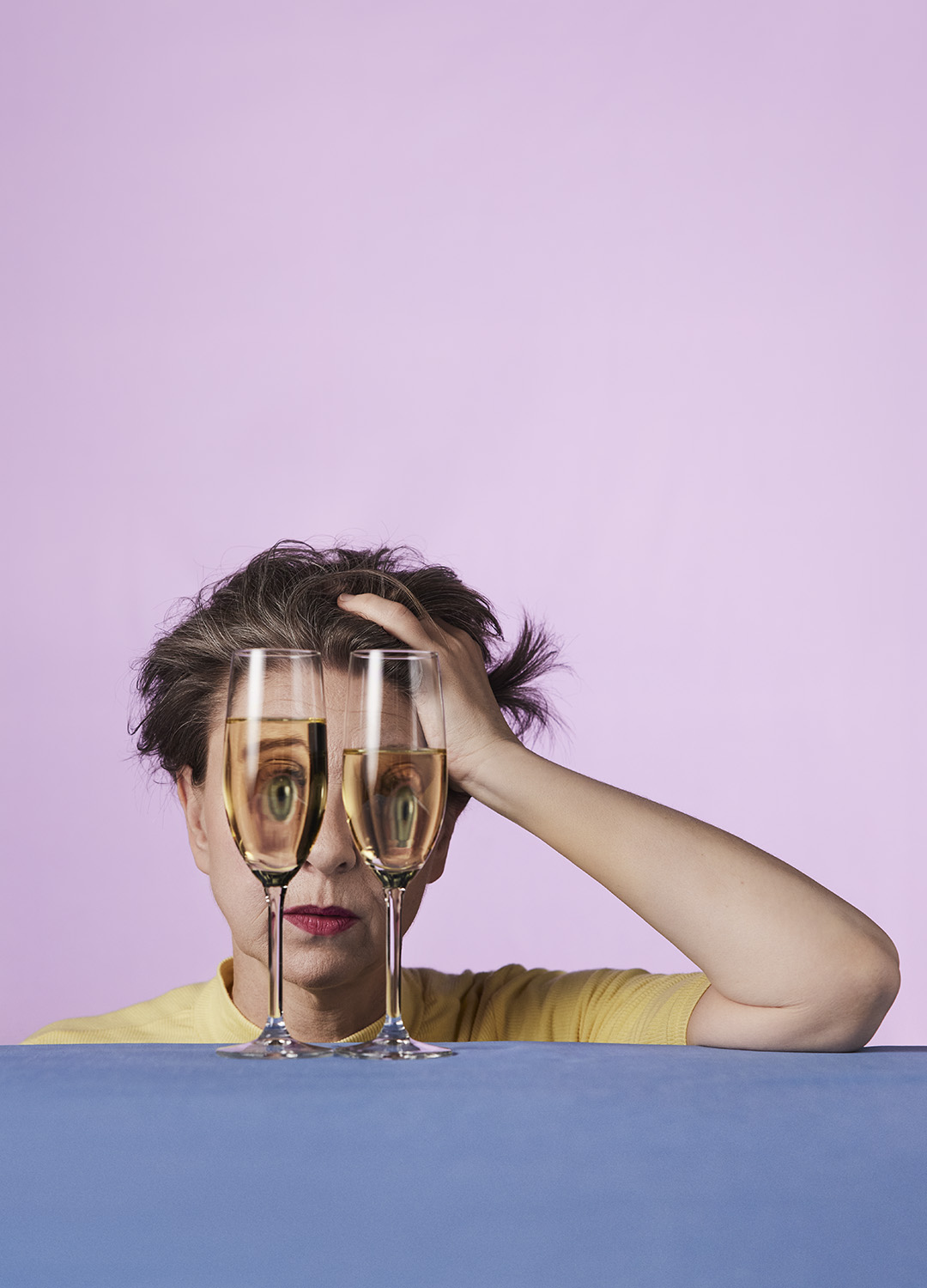 Kvinna sitter vid ett bord med huvudet lutat i handen med två vinglas framför ansiktet som gör att ögonen ser komiskt förvridna ut genom. 
