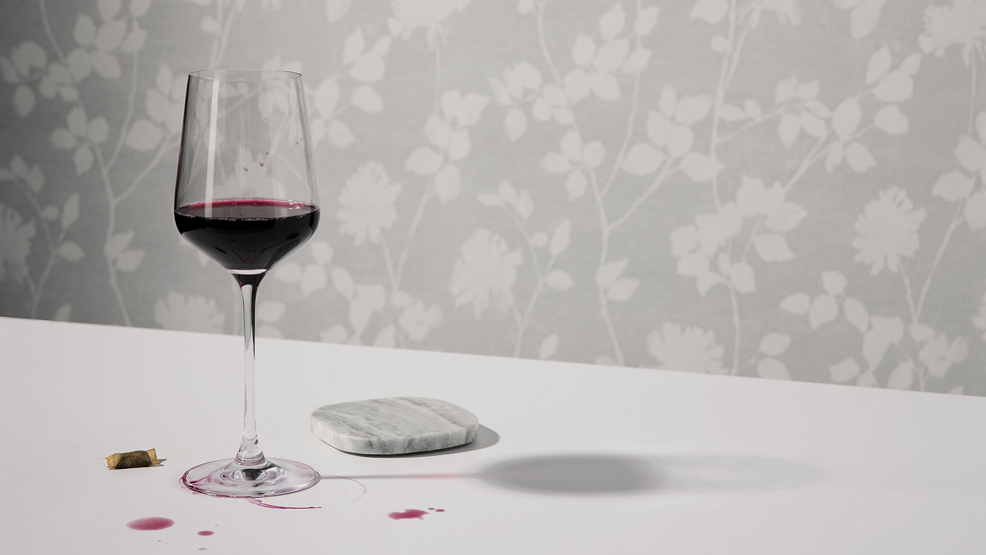 Vinglas till hälften fyllt med rödvin jämte ett glasunderlägg på ett vitt bord med rödvinsspill och en använd snus. 