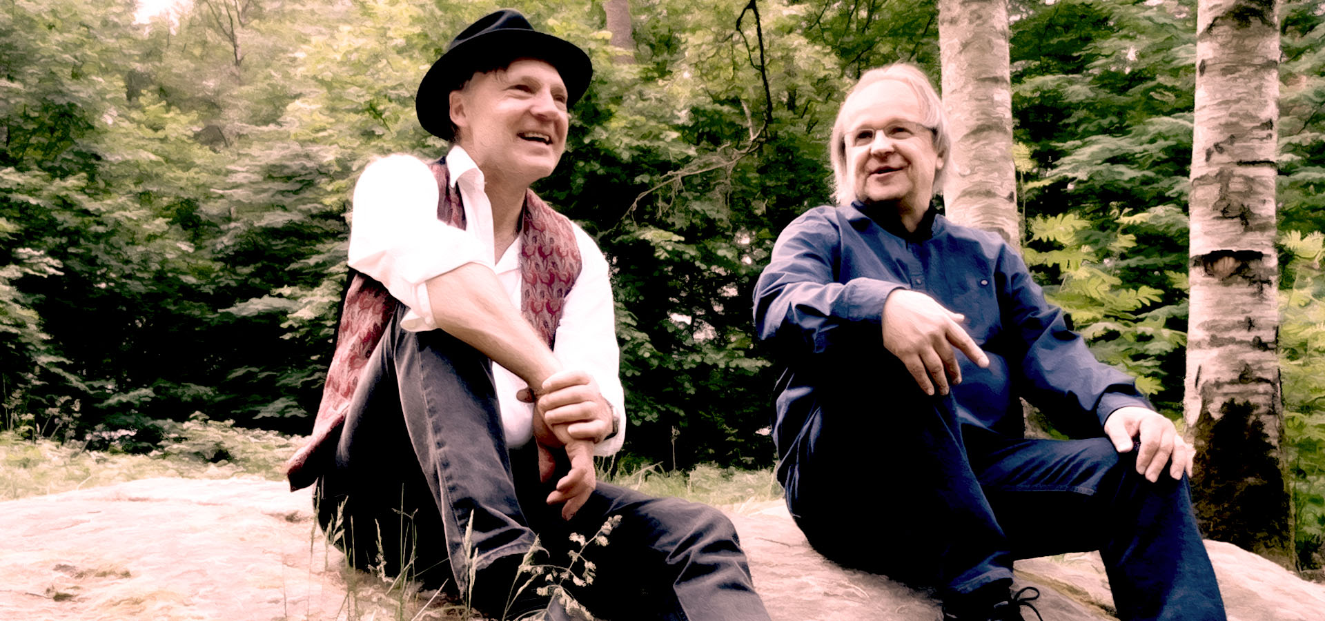 Artisterna Lennart och Markku sitter avslappnat på en stenhäll med skog och gröna blad i bakgrunden. Lennart bär hatt och väst, Markku i mörkblå skjorta. 