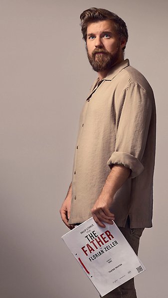 En person står i profil mot ljus bakgrund och håller en bok med titeln “Philip Lithner spelar The Father av Florian Zeller”.